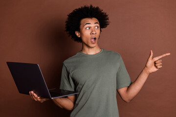 Photo portrait of young guy in gray t shirt remote holding netbook point finger mockup surprised isolated on brown color background