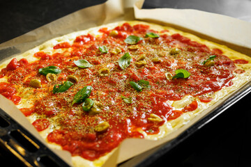 A vibrant, uncooked pizza with tomato sauce, mozzarella cheese, basil leaves, and garlic on a baking tray.