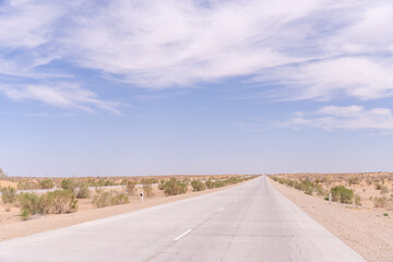 A long, empty road with a clear blue sky above. The road is surrounded by a desert landscape