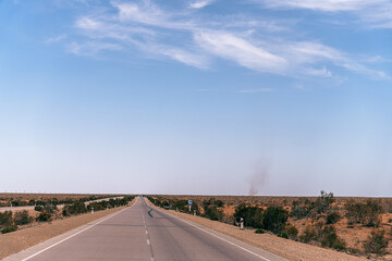 A long, empty road with a blue sky above. The road is surrounded by a desert landscape
