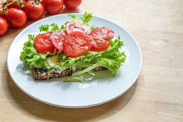 Healthy sandwich, dark whole meal bread with vegan cream cheese, lettuce and tomatoes, seasoned with pepper and salt on a white plate and a wooden table, copy space