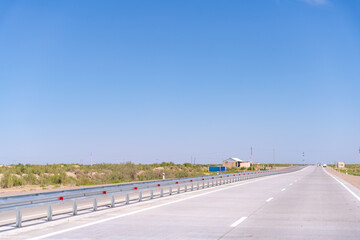 A long, empty highway with a blue sky above. The road is lined with red and white guardrails