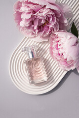 A bottle of women's perfume lies on a white podium in the form of an arch with delicate lilac peonies. top view. The concept of beauty and perfume.