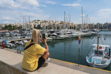 A young girl tourist takes a photo on her phone in Athens, Greece, in the yacht port of Pasalimani on a summer day.