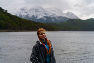 Beautiful young woman with red hair a smiling jacket, with the Beagle Channel and the snow-capped Andean mountains of Tierra del Fuego National Park, Ushuaia - Argentina.