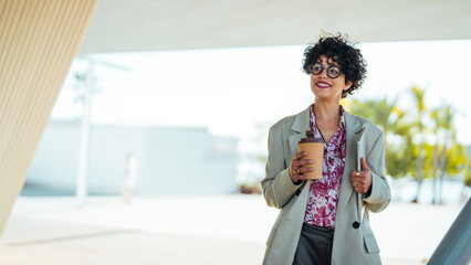 A joyful Hispanic businesswoman strolls outdoors, sipping coffee and giving a thumbs up, dressed in...