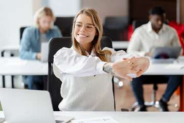 Young, smiling businesswoman stretching arms at office desk, break