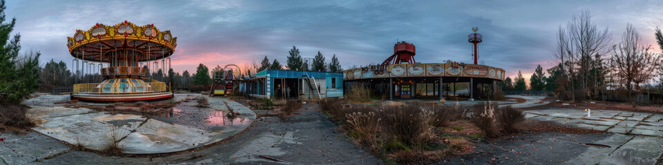 An abandoned amusement park in Pripyat, Ukraine, stands silent at dusk. A carousel, a roller coaster, and other rides are visible in the foreground, covered in dust and overgrown with vegetation