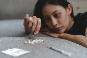 drug addict asian young woman hand holding pills, medicament with narcotic syringe on sofa at home, abuse overdose