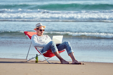 digital nomad mature hispanic man sitting on the shore of the beach working with his laptop