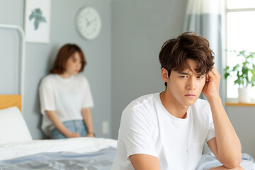 ED TREATMENT (Erectile Dysfunction). A man in a white t-shirt is sitting on the bed, holding his head with one hand and looking sad while another woman stands behind him from far away in a bedroom 