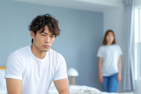 ED TREATMENT (Erectile Dysfunction). A man in a white t-shirt is sitting on the bed, holding his head with one hand and looking sad while another woman stands behind him from far away in a bedroom 