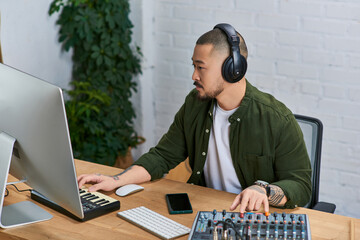A handsome Asian man, wearing headphones, works on a musical project in his studio.