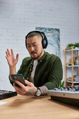 A man in casual attire sits in his studio, headphones on, holding a phone while looking at the...