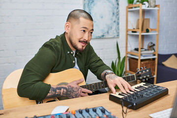 A handsome Asian man plays the guitar and MIDI keyboard while creating music in his studio.