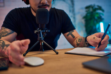 An Asian man in a studio, recording a podcast, with a microphone in front of him and a notebook and...