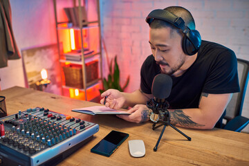 A man records a podcast in his home studio, with a microphone, audio mixer, and notebook in front of him.