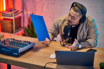 A handsome Asian man sits in his home studio, headphones on, recording a podcast with a microphone.