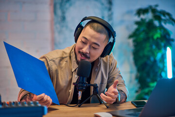 A handsome Asian man in headphones is recording a podcast in his studio, holding a blue sheet of...