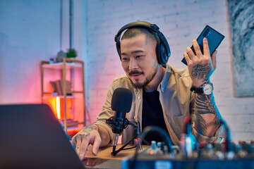 An Asian man in headphones speaks into a microphone while recording a podcast.