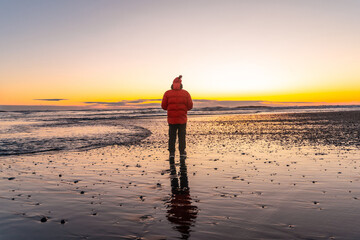 A man stands on a beach at sunset, wearing a red jacket © unai