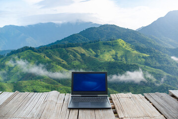A laptop placed on a wooden deck with a breathtaking view of lush green mountains and misty clouds in the background.