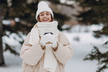 Happy smiling young woman with coffee hot drink cup enjoys winter weather at snowy winter park