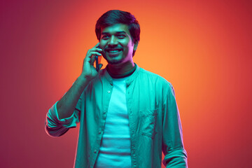 Smiling young Indian man looking at distance and talking on phone in neon light against gradient studio background. Concept of fashion and style, human emotions, communication, cellular connection.