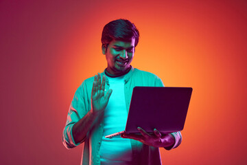 Young, confident Indian man dressed casual outfit holds meeting online using laptop in neon light against gradient studio background. Concept of fashion and style, human emotions, study, work online.