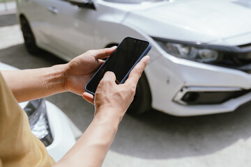 A young adult man, holding a smartphone, takes a photo of his dented car after an accident. An...