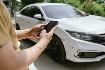 A young adult man, holding a smartphone, takes a photo of his dented car after an accident. An insurance agent with a clipboard examines the damage, preparing a claim report.