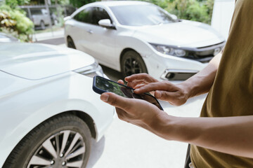 A young adult man, holding a smartphone, takes a photo of his dented car after an accident. An...