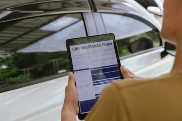 A young adult man, holding a smartphone, takes a photo of his dented car after an accident. An...