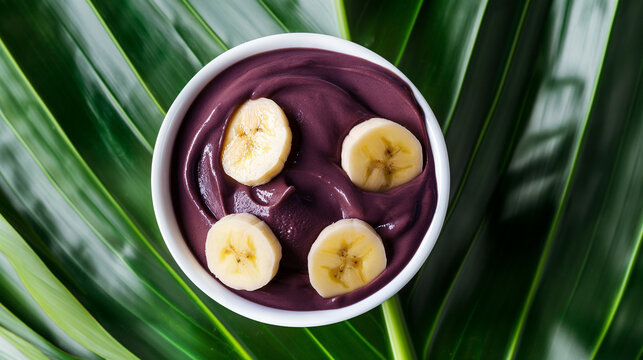 Uma pequena tigela branca de a&ccedil;a&iacute; brasileiro com fatias de banana por cima, colocada contra um fundo de folhas de palmeira verdes em uma vista superior