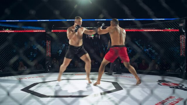 Caucasian MMA Fighters Exchange Powerful Punches and Kicks in an Octagon During a Championship Battle. Referee Supervising the Fight, Audience Watching Behind the Ring. Footage From Behind a Fence