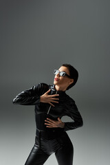A woman in black leather jumpsuit poses against a grey backdrop.