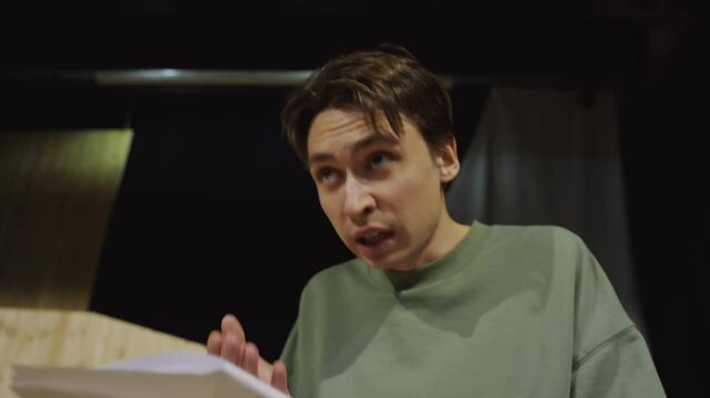 Young male actor reading monologue on paper and performing with expression during rehearsal on theatre stage