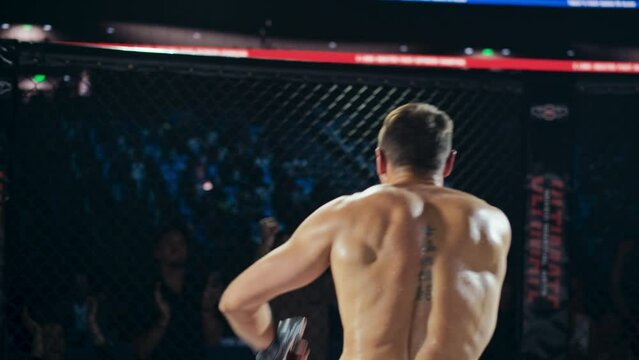 Referee Declares the Winner in a Tough MMA Bout Between Two Determined Male Fighters. Winning Fighter Raising His Hands in Triumph, Running and Climbing the Octagon Fence to Greet the Crowd