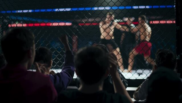 Excited Audience Watching Two Male Mixed Martial Artists Fight Inside an Octagon Cage, Using Powerful Kicks and Punches. Referee in Charge Watching the Match, Crowd is Cheering