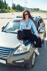 A girl sits on the hood of a car on a sunny summer day.