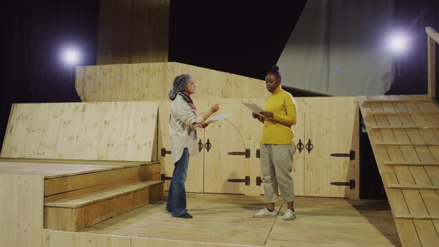 Young African American actress holding scrip on paper, listening to female theatre director and performing on stage during rehearsal