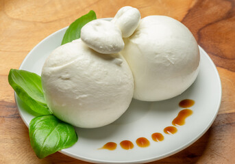 Ball of mozzarella or burrata cheese with fresh basil leaves on white plate on wooden background.