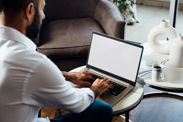 Over shoulder view of a young business man using computer laptop in front of an blank white computer screen in his home living room. Photo ready for mockup.