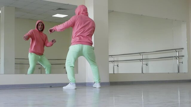 A young stylish guy looks into the reflection of the mirror and dances. A man in a hoodie dances a regular street dance in a studio.
