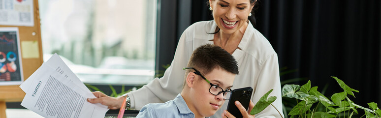 A mother smiles at her son as they work side-by-side in a modern office, showcasing inclusion and...