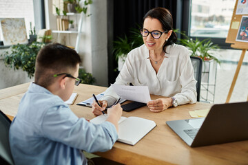 A mother and her son with Down syndrome work together at a desk in an office, showcasing inclusion...