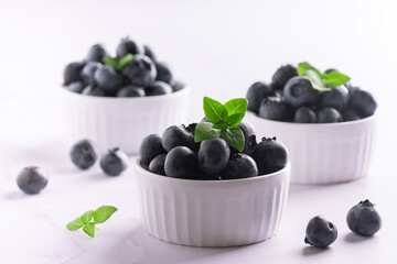 Tasty aromatic blueberries in white ceramic bowls on light marble background close up.