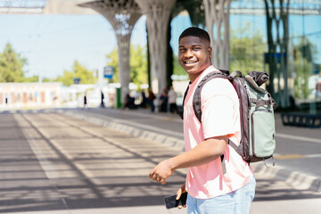 African man wearing a pink shirt and a green backpack is smiling and holding a cell phone. He is walking down the street, possibly heading to a destination.