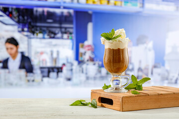 Iced coffee in a glass cup with cream and fresh mint on a wooden board on bar counter tabletop blurred restaurant bar background