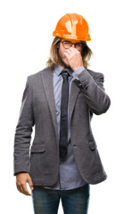 Young handsome architec man with long hair wearing safety helmet over isolated background smelling something stinky and disgusting, intolerable smell, holding breath with fingers on nose. Bad smells